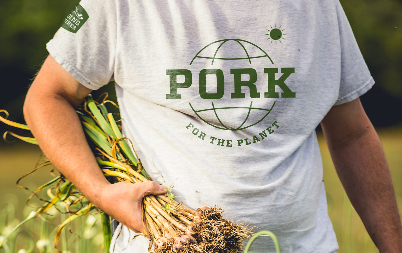 A close up of a person wearing a Singing Pastures "pork the planet" t-shirt and carrying stalks of freshly picked garlic.