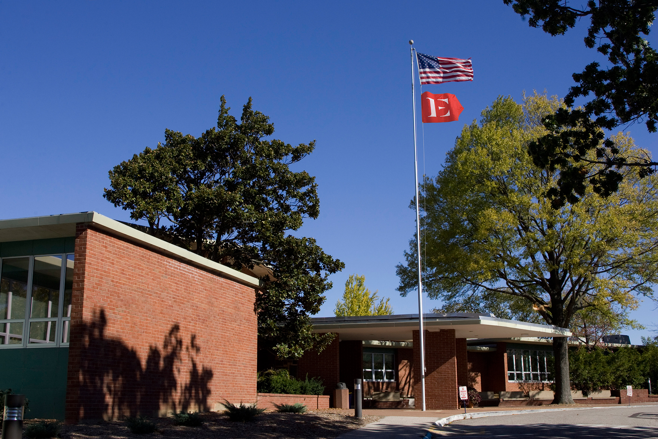 The Expeditionary Learning "capital E in a crimson chevron" logo on a flag flying in front of a suburban school.
