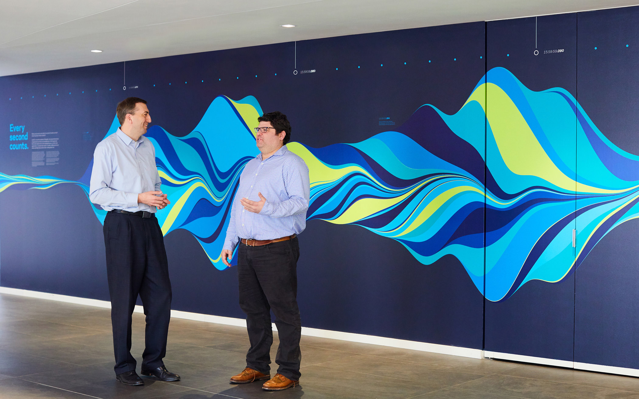 Two KCG employees stand in front of a multi-colored wave pattern mural that depicts one second of market trading activity.