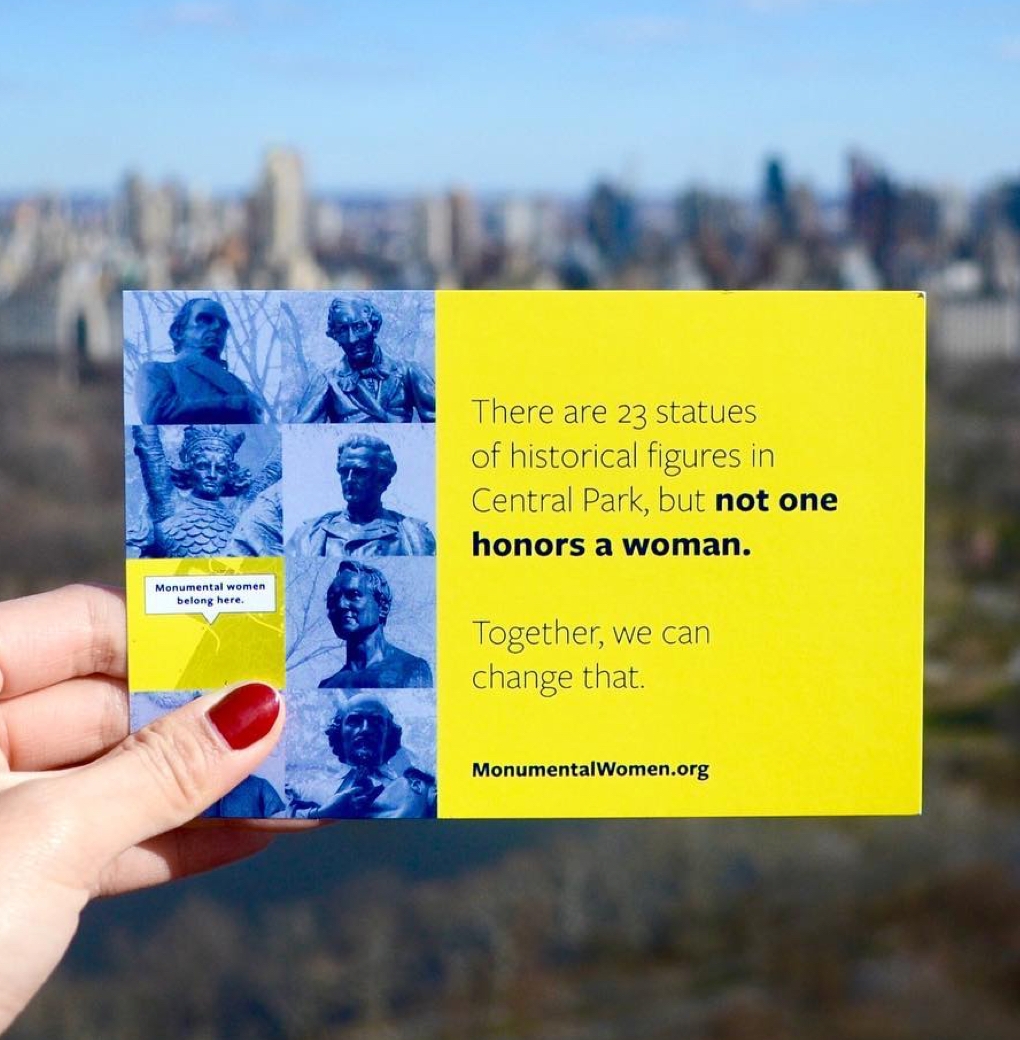 A hand holds a yellow Monumental Womand postcard aloft with Central Park out of focus in the background. 