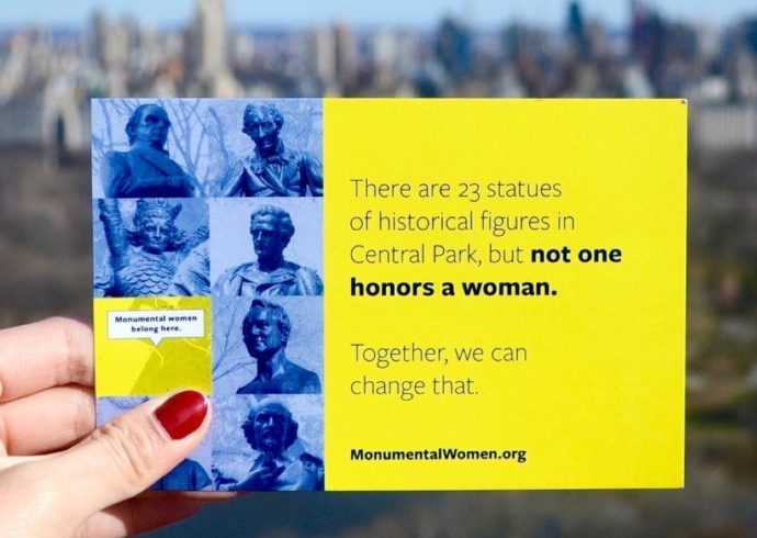 A hand holds a yellow Monumental Womand postcard aloft with Central Park out of focus in the background.