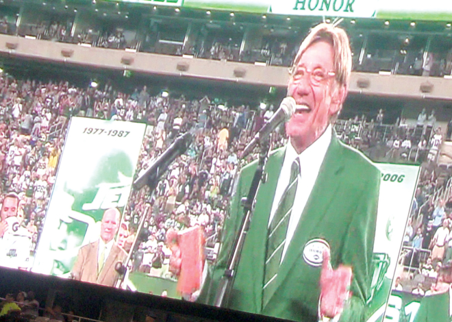 Joe Namath flanked by other New York Jets veterans in green jackets making an announcement on the football field.