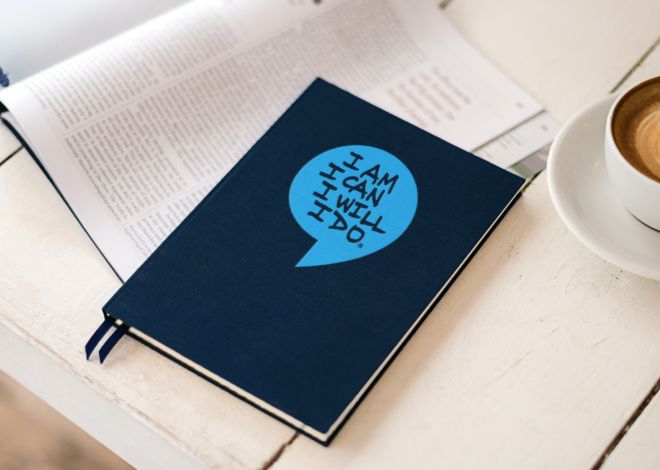 A navy blue journal with a bright blue enlarged speech bubble with the words, “I am I can I will I do” in blue, capitalized letters on a desk.