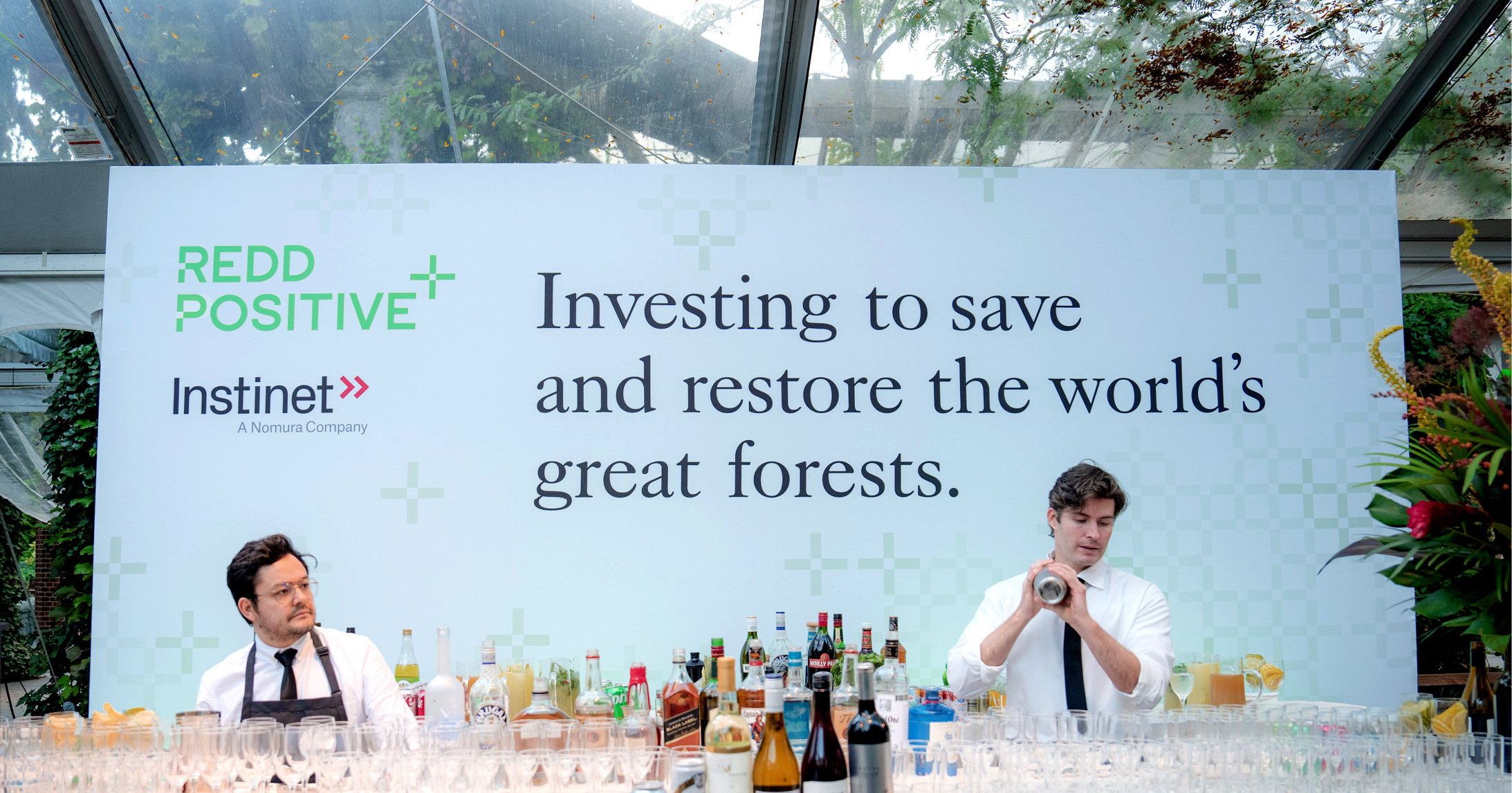 Two Redd Positive Cocktails and Conservation launch event food and drink servers in front of a large banner featuring the Redd Positive and Instinet logos with a large headline that reads, Investing to save and restore the world’s great forests. 